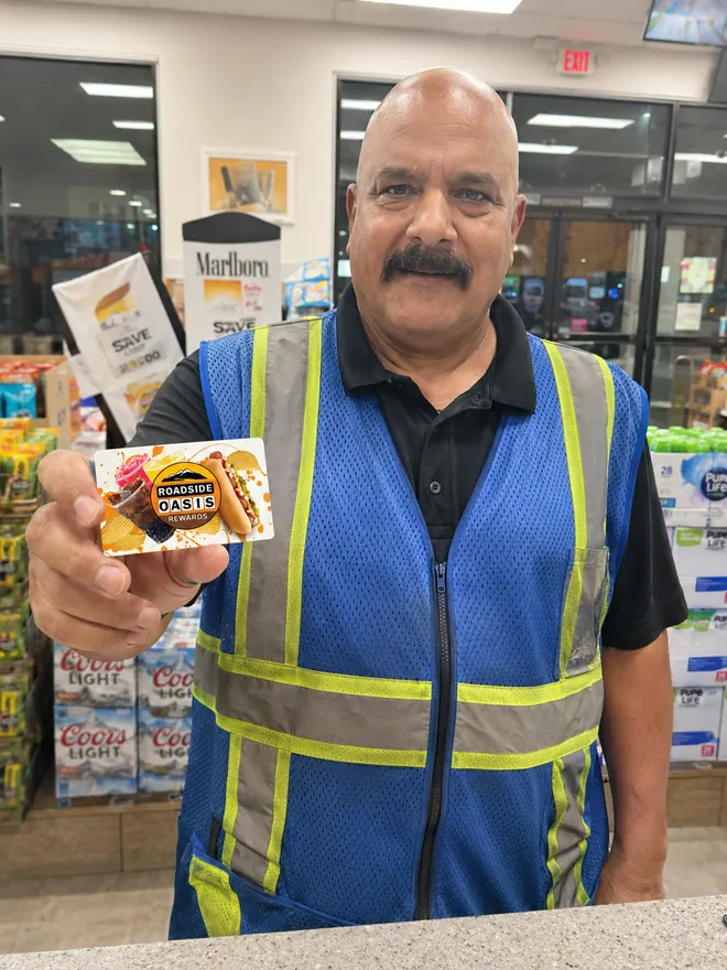 Roadside Oasis employee in a blue hi-vis vest holding a Rewards card inside the convenience store