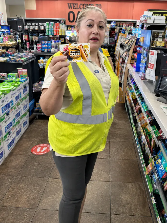 Store associate in a yellow hi-vis vest holding a Roadside Oasis Rewards card in the snack aisle