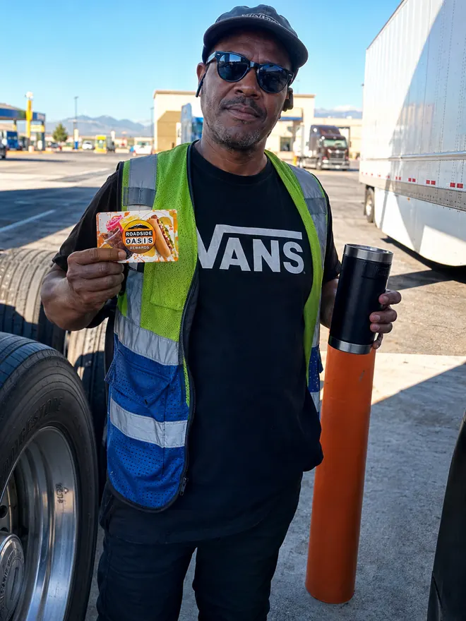Truck driver in sunglasses and a safety vest holding his Rewards card beside his trailer at the fuel island
